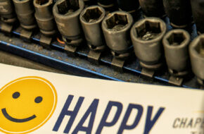 photo of a set of wrenches beside a bumper sticker with a yellow smiley face that reads "Drive Happy" and a blue ball cap with a yellow smiley face