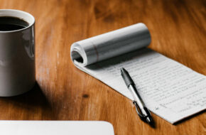 Coffee in a white mug on table with laptop and a notepad beside it
