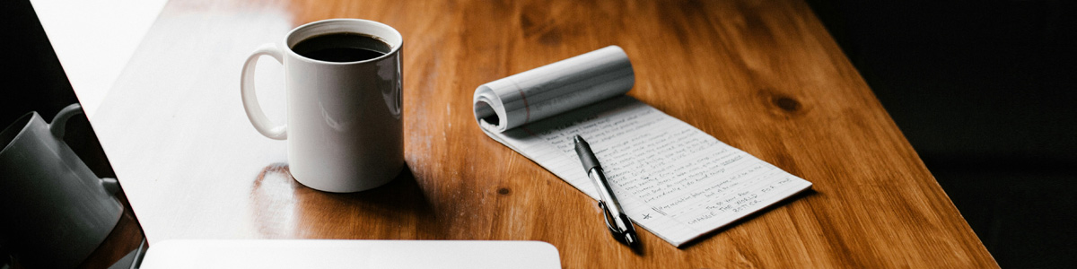 Coffee in a white mug on table with laptop and a notepad beside it